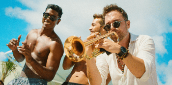 Three men outside by a pool with one playing the trumpet
