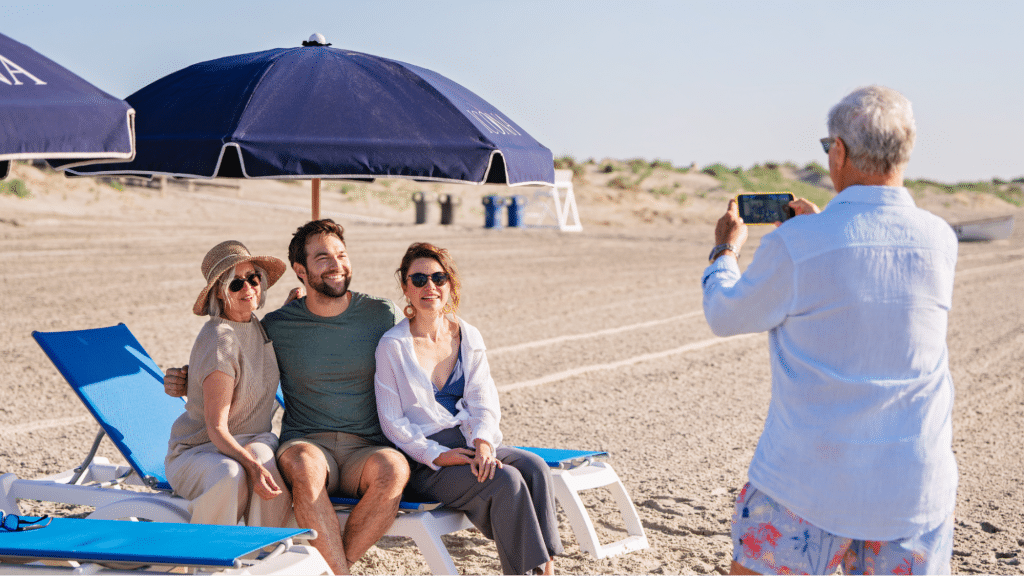 Three travelers are posing for a photo on a beach resort. They are sitting on a blue beach chair with a blue umbrella blocking the sun.