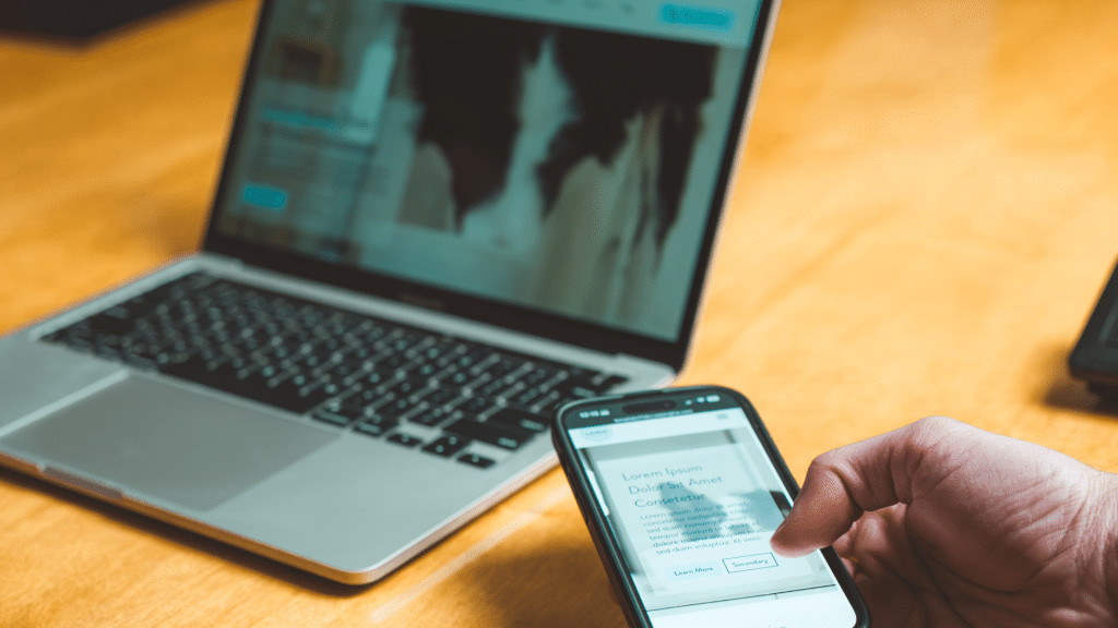 A laptop is sitting open on a table while a hand holding a cell phone is visible next to it.